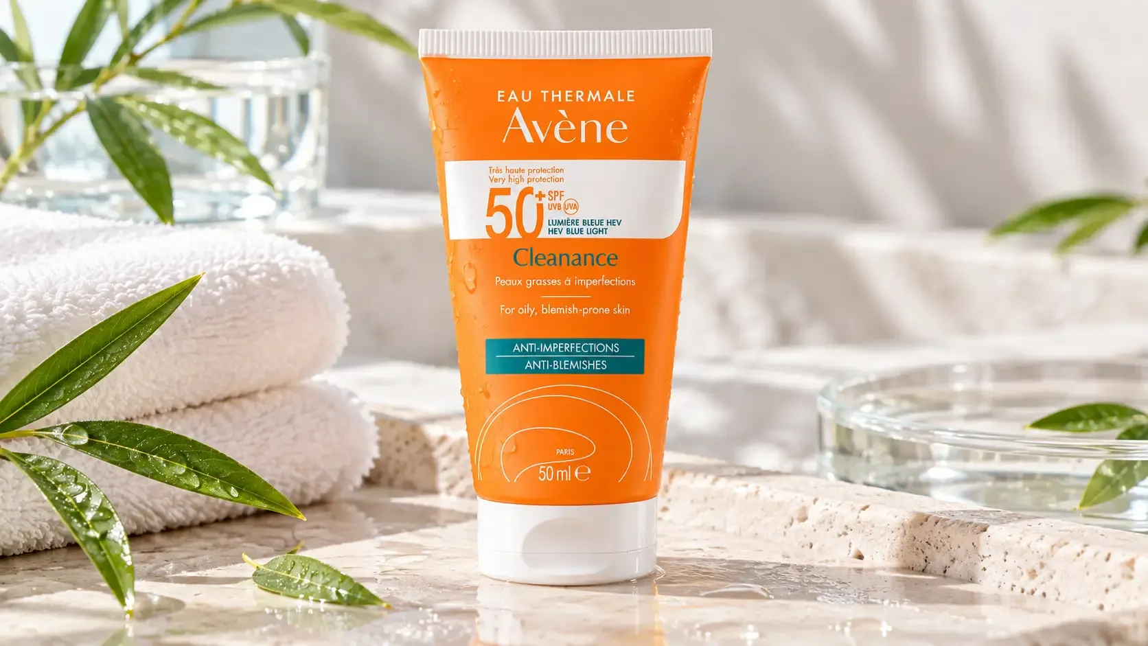 أفين كلينانس واقي شمس SPF 50+ بحجم 50 مل في مشهد عناية بالبشرة مع مناشف بيضاء وأوراق خضراء ومياه منعشة